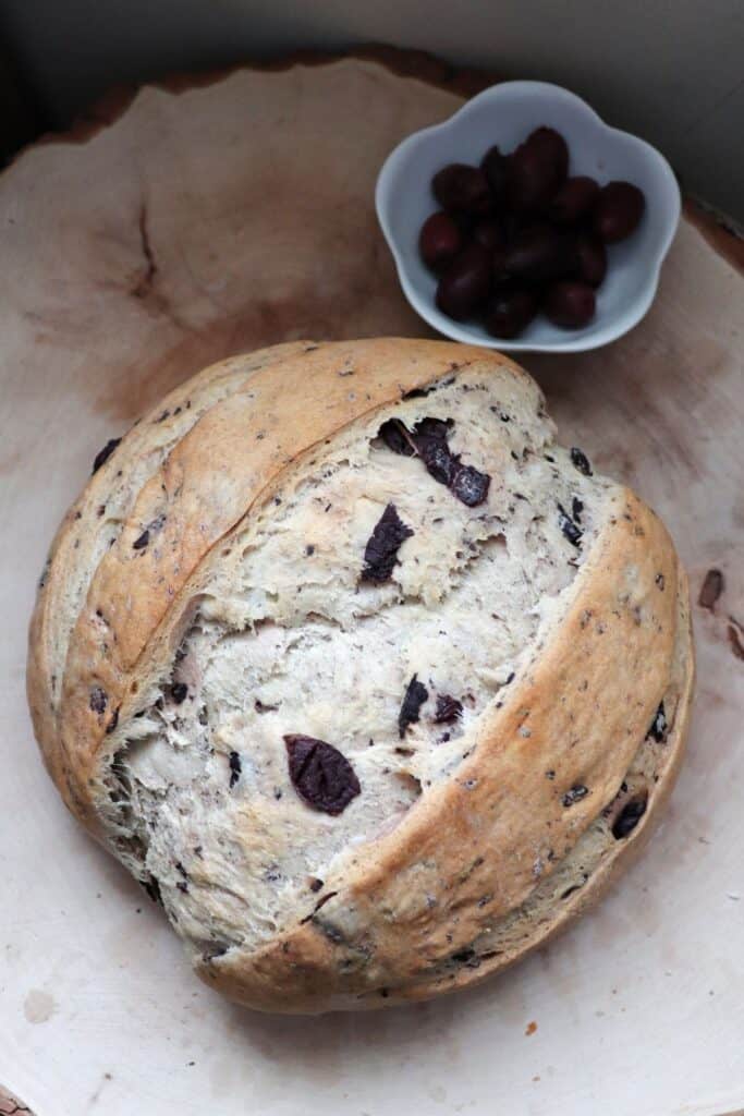 A loaf of kalamata olive bread as seen from above sitting on a wood board with a bowl of olives sitting next to it. 