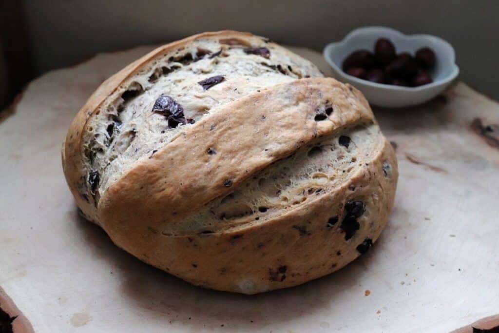 A loaf of kalamata olive bread on a wood board with a bowl of olives in the background. 