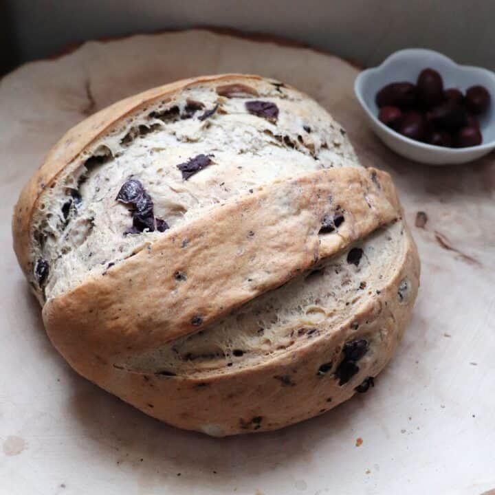 A round loaf of kalamata olive bread on a board sitting next to a bowl of olives.