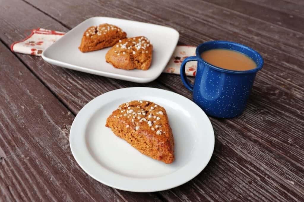 A white plate with a gingerbread scone sitting on it, a blue cup full of tea next to it, a square plate with 2 scones on it in the background.