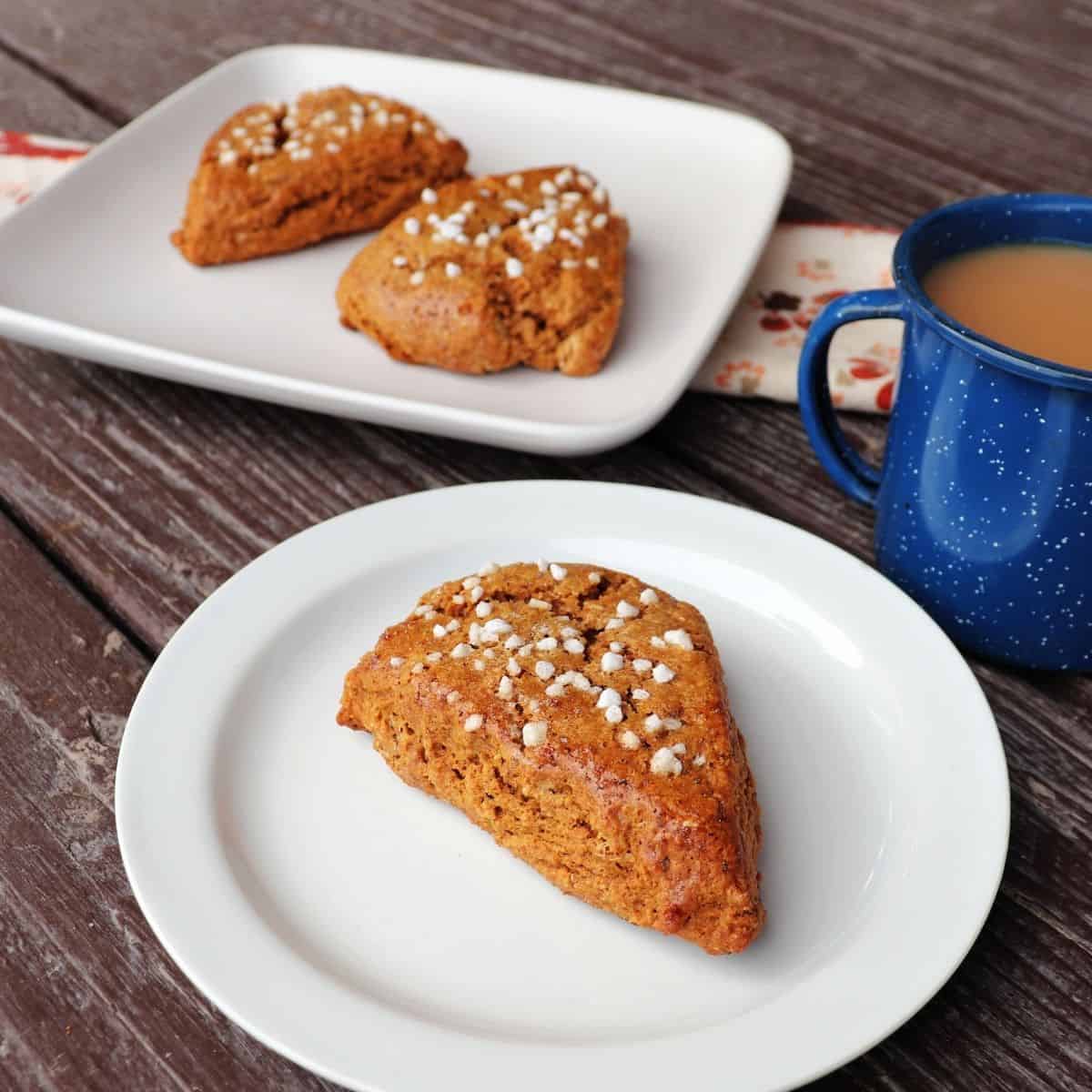 A white plate with a gingerbread scone sitting on it, a blue cup full of tea next to it, a square plate with 2 scones on it in the background.