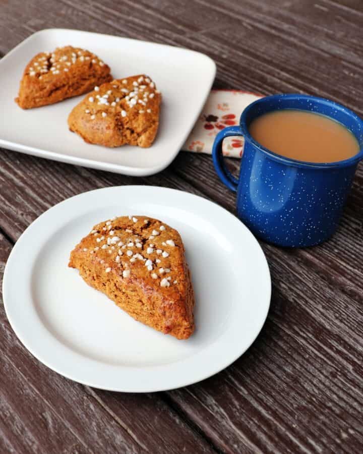 A white plate with a gingerbread scone sitting on it, a blue cup full of tea next to it, a square plate with 2 scones on it in the background.
