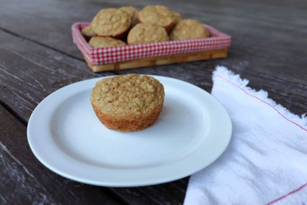 An applesauce bran muffin on a white plate with a white napkin sitting next to you. A basket with more muffins sits in the background.