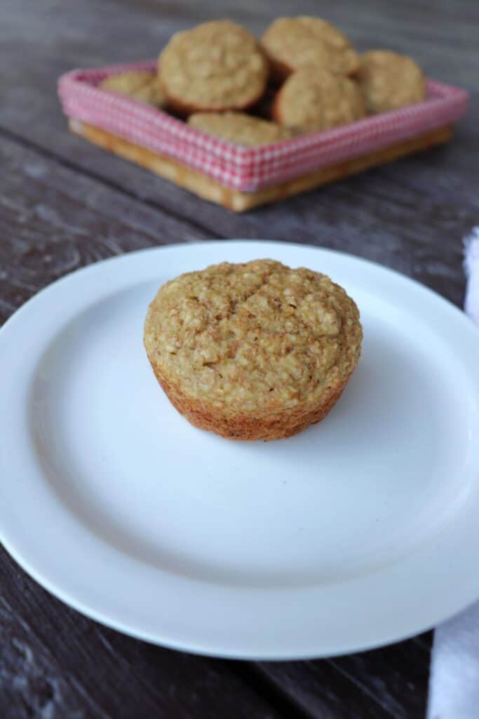An applesauce bran muffin on a white plate with a basket of more muffins in the background.