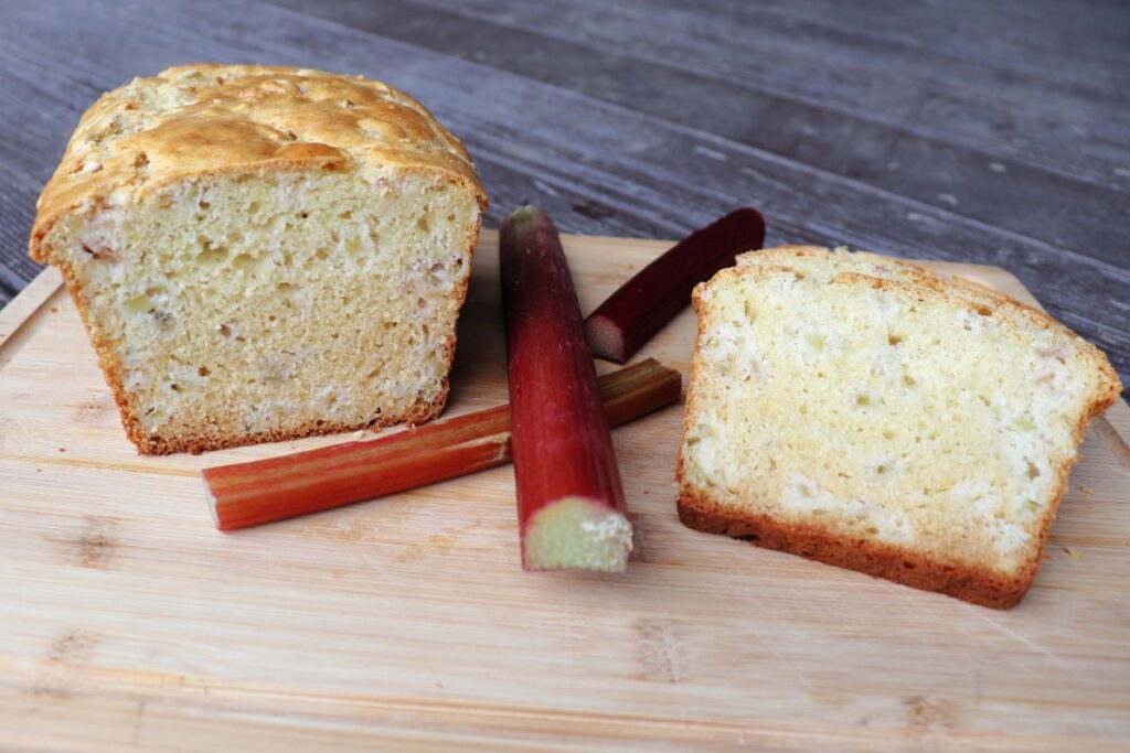 Slices of rhubarb board to the right of a wood cutting board and the remaining loaf on the left. Stalks of red rhubarb between.