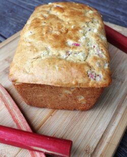 A loaf of rhubarb bread on a board surrounded by fresh red stalks of rhubarb.