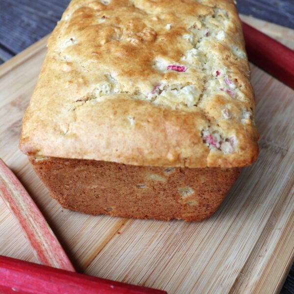 A loaf of rhubarb bread on a board surrounded by fresh red stalks of rhubarb.