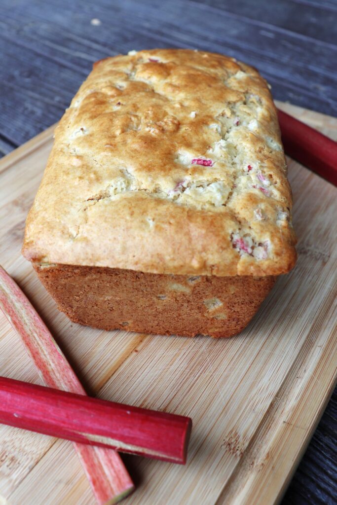 A loaf of rhubarb bread on a board with stalks of red rhubarb around it.