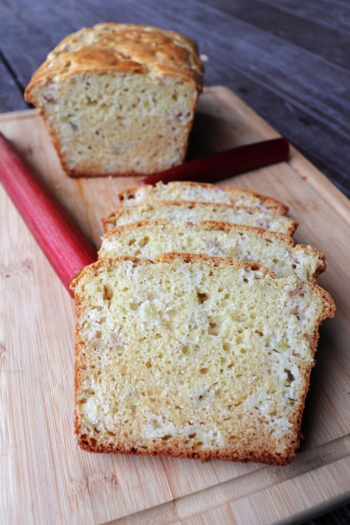 Slices of rhubarb bread on a board with fresh stalks of rhubarb and the remaining loaf in the background.