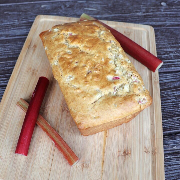 A loaf of buttermilk rhubarb bread on a cutting board with fresh stalks of rhubarb surrounding it.