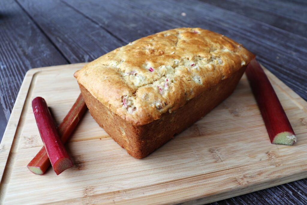 A loaf of rhubarb bread on a board surrounded by fresh red rhubarb stalks.