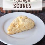 A cheddar cheese scone on a white plate with more scones on a cutting board in the background. with text overlay reading cheese scones.