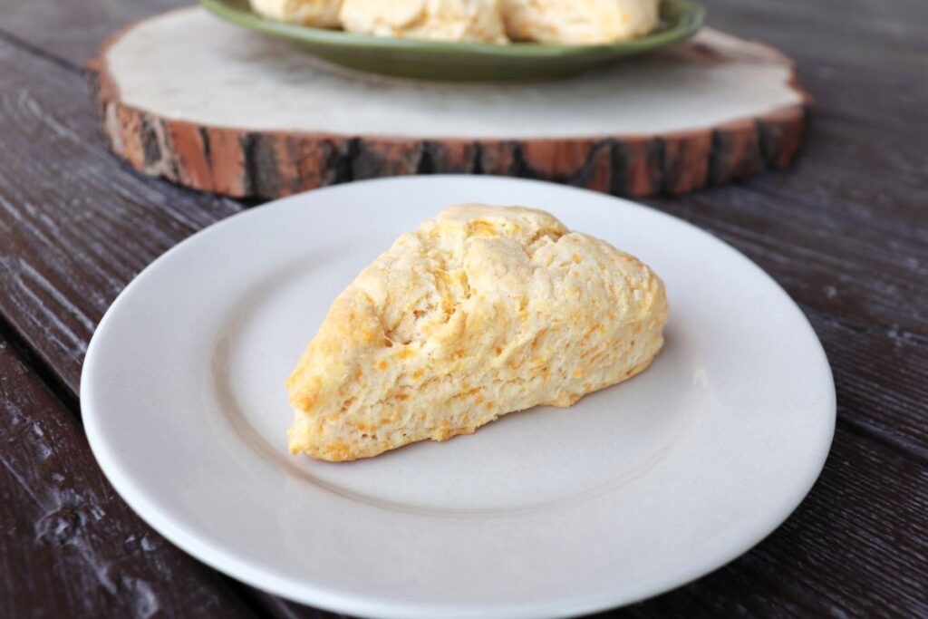 A cheddar cheese scone on a white plate with more scones on a cutting board in the background.