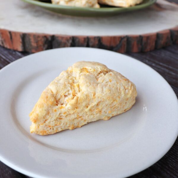 A cheddar cheese scone on a white plate with more scones on a cutting board in the background.
