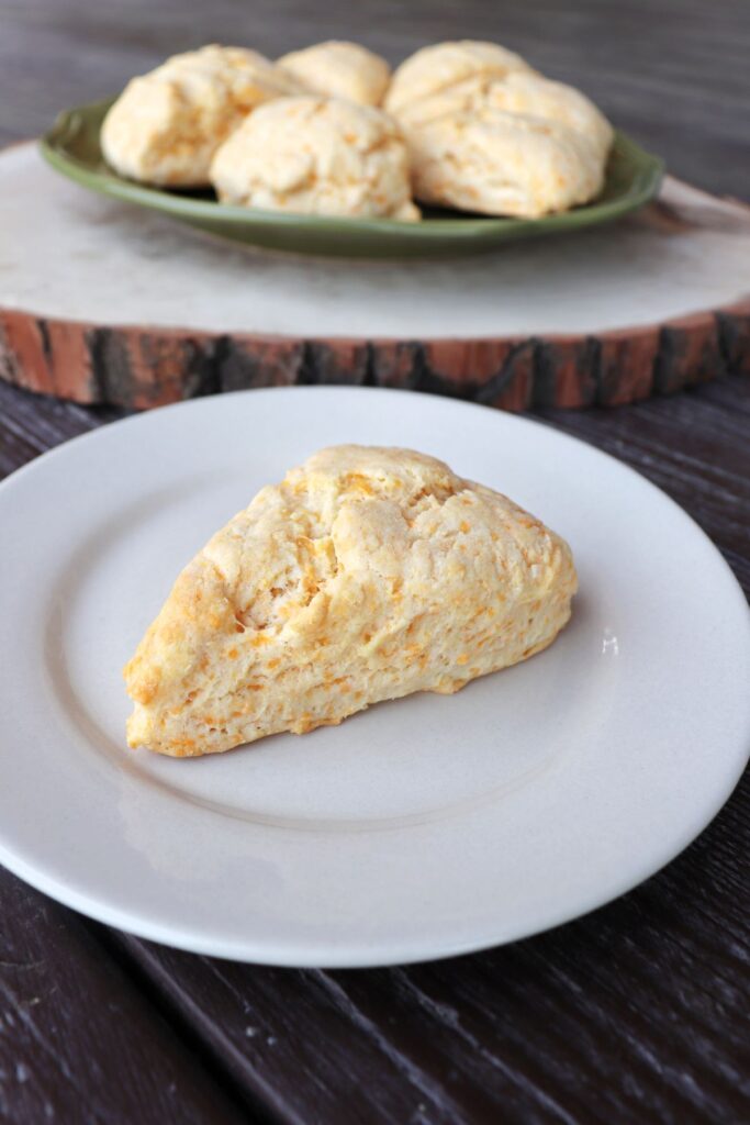 A cheddar cheese scone on a white plate with more scones on a cutting board in the background.