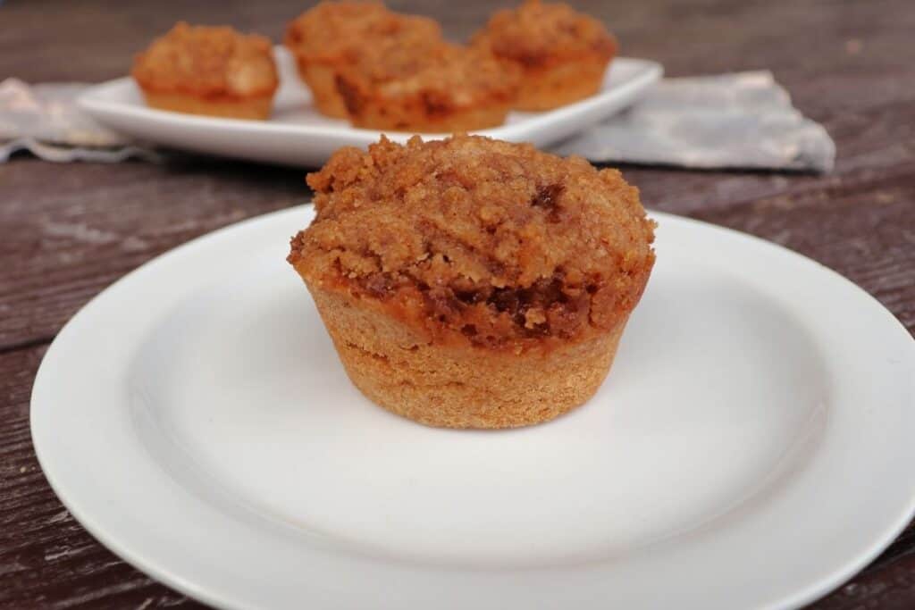 A cinnamon muffin on a white plate with a platter of more muffins in the background.