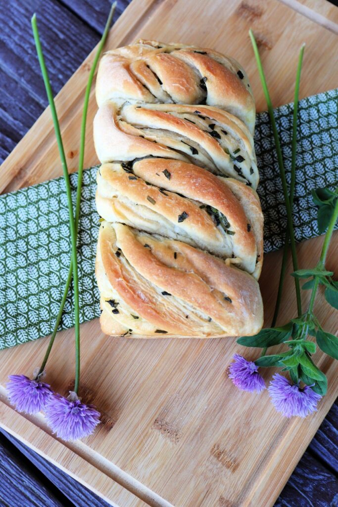 A golden twisted loaf of herb bread as seen from above sits on a green cloth on top of a wooden board surrounded by fresh herbs.