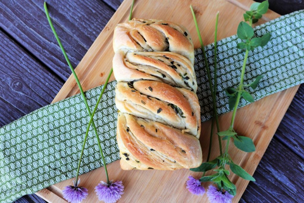 A golden twisted loaf of herb bread as seen from above sits on a green cloth on top of a wooden board surrounded by fresh herbs.