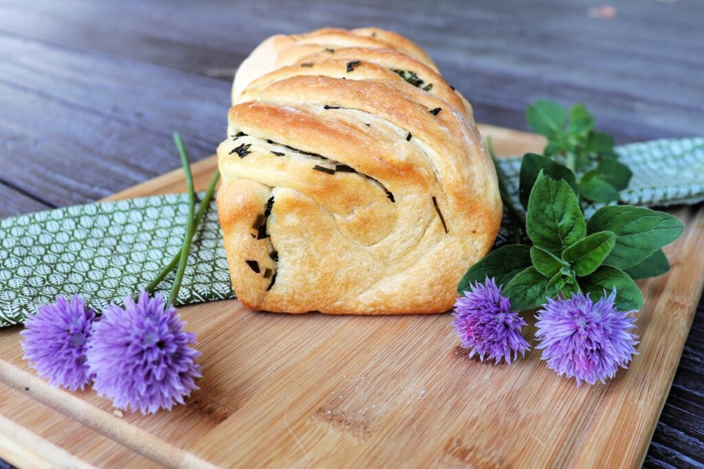 A loaf of fresh herb bread seen from the end with bits of herbs exposed in the crust sits on a green cloth on top of a board. Fresh herbs surround the loaf.