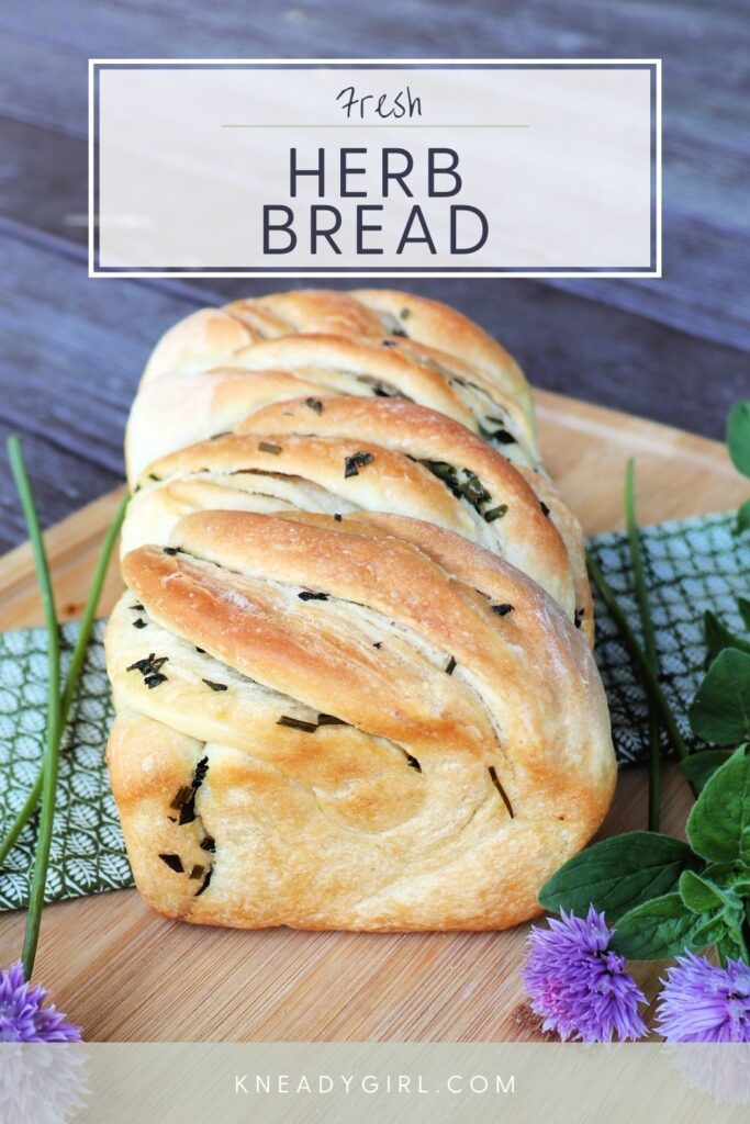 A golden loaf of bread with a twisted top and bits of green herbs showing, sitting on a cloth surrounded by green herbs. A text overlay reads fresh herb bread. 