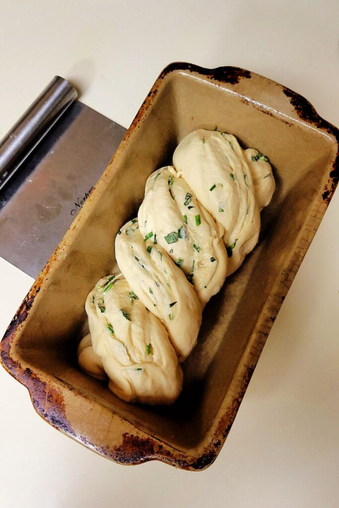 Twisted raw bread dough with exposed layers of green herbs sitting in a brown bread pan.