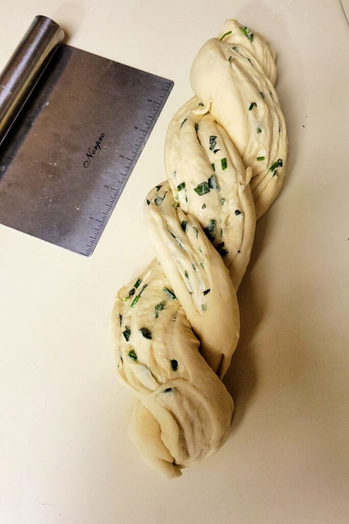 Two halves of raw bread dough twisted together to form one loaf with layers of herbs exposed.