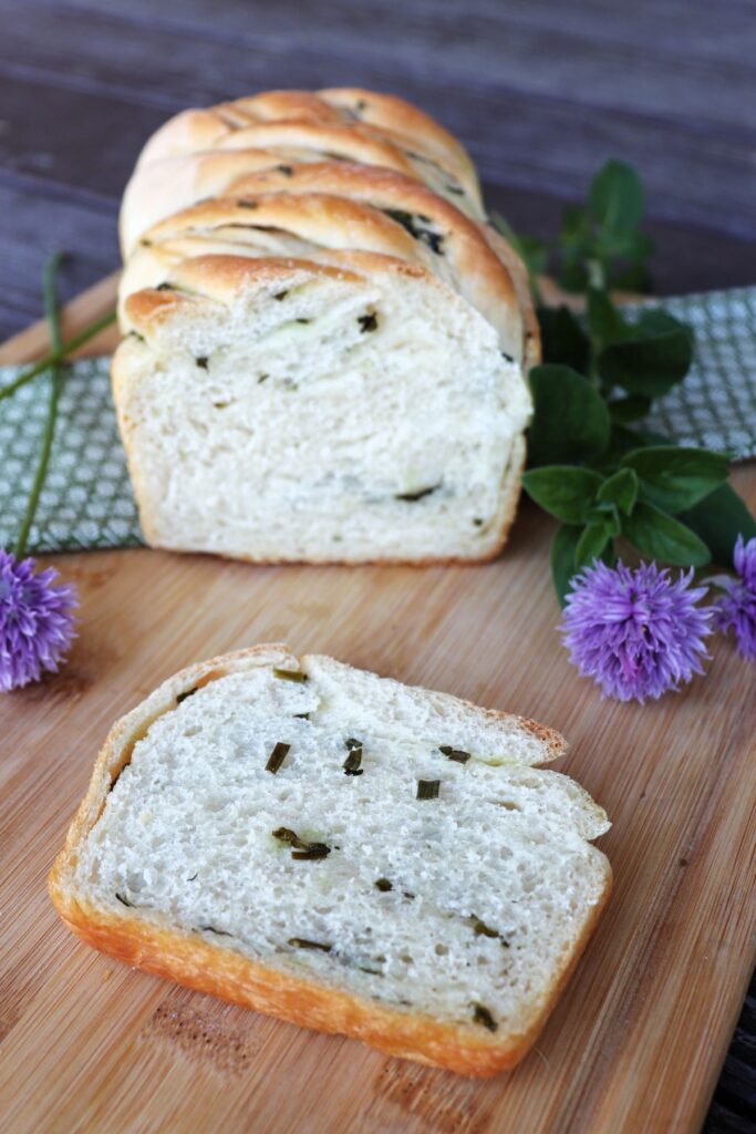 A slice of herb bread sitting on a board with bits of herbs exposed, behind it sits the remaining loaf surrounded by fresh herbs.