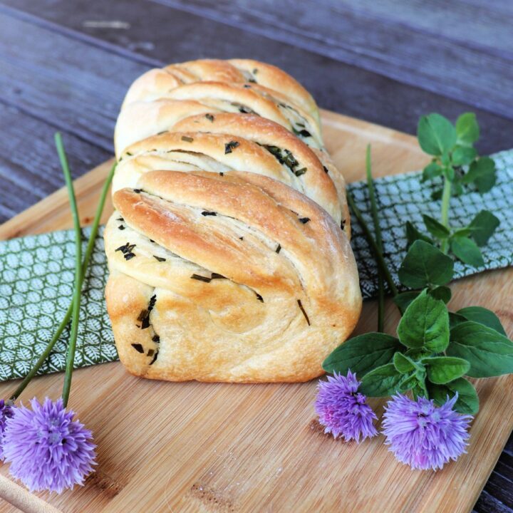 A loaf of fresh herb bread sits on a board surrounded by fresh herbs.