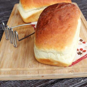 2 loaves of mashed potato bread sitting on a board with a metal potato masher between them.