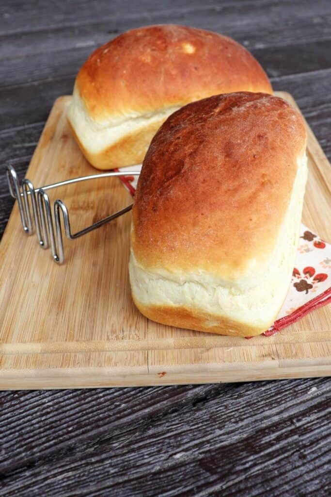 Two loaves of mashed potato bread on a board with metal potato masher in between.