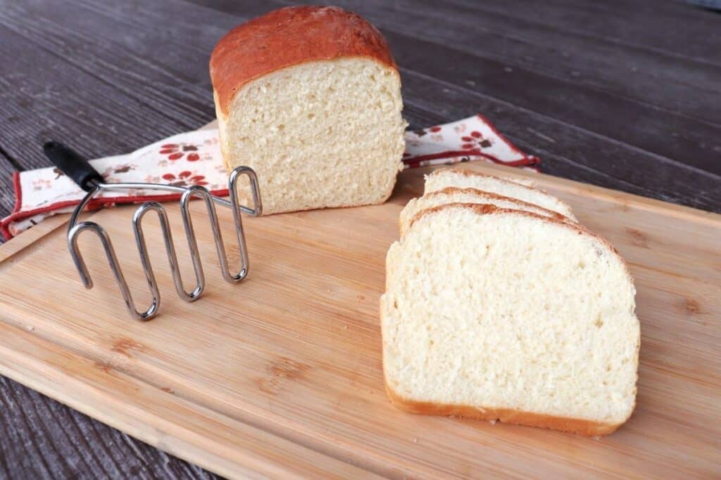 Slices of mashed potato bread on a board with remaining loaf and metal potato masher in the background.