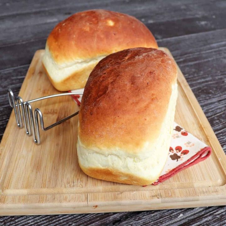 2 loaves of mashed potato bread on a board with a metal potato masher between them.