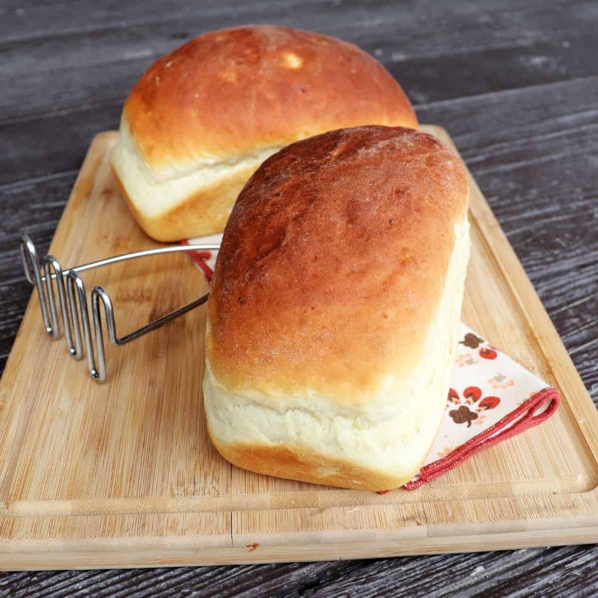 2 loaves of mashed potato bread on a board with a metal potato masher between them.