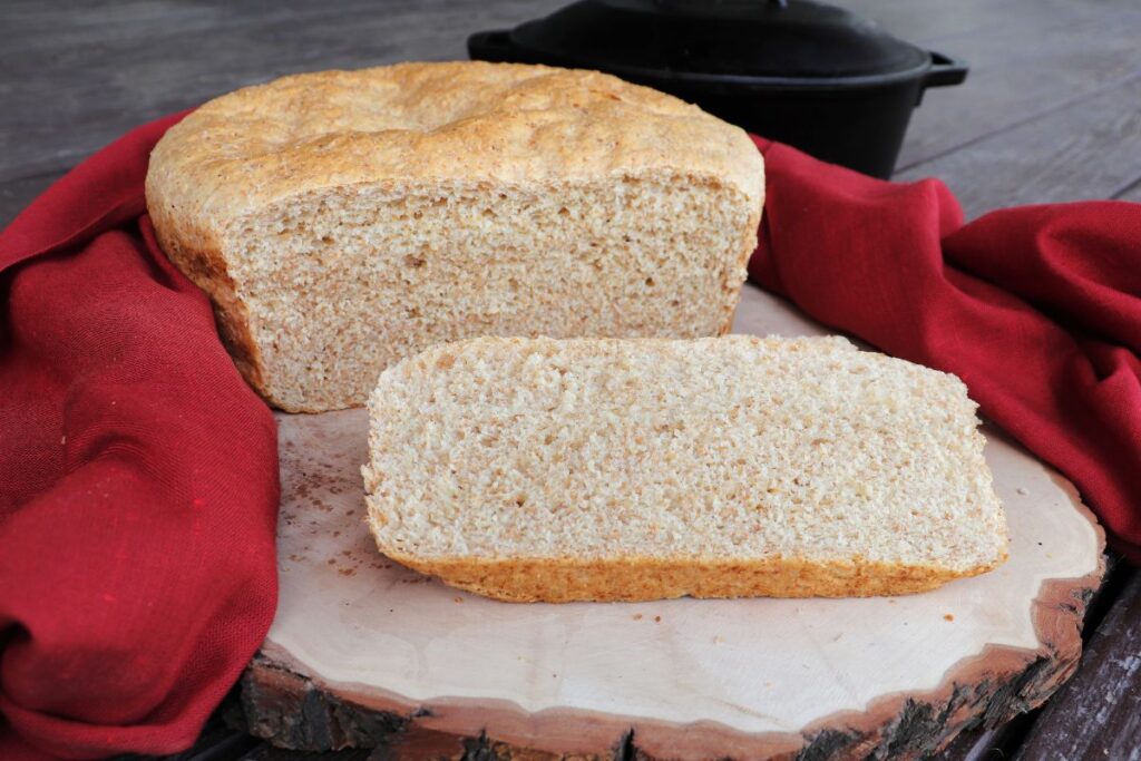 Slices of shepherd's bread sitting on a board with remaining loaf behind it. The bread is surrounded by a red cloth and a black Dutch oven sits in the background.