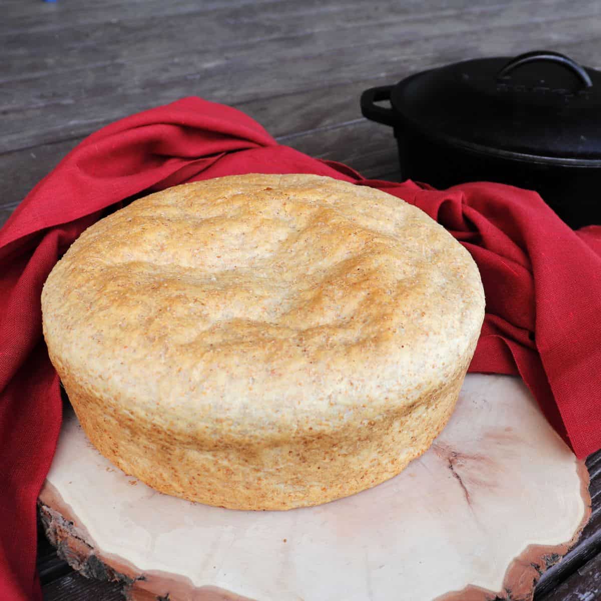 A loaf of shepherd's bread sitting on a board with a red cloth and a dutch oven in the background.