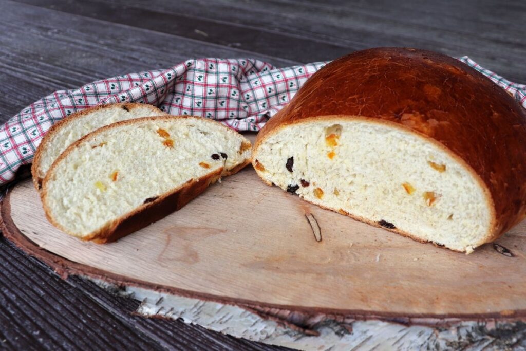 Slices of julekake bread sitting on a board next to the remaining loaf with bits of dried fruit exposed in the bread. A cloth sits behind it.