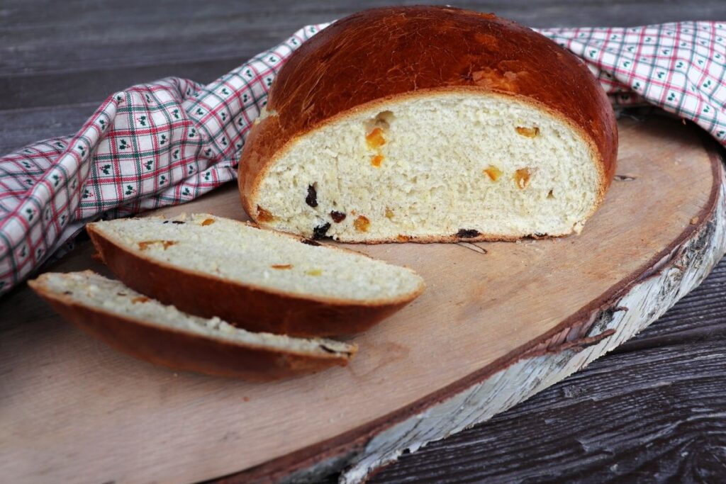 Slices of julekake bread sitting on a board next to the remaining loaf with bits of dried fruit exposed in the bread. A cloth sits behind it.