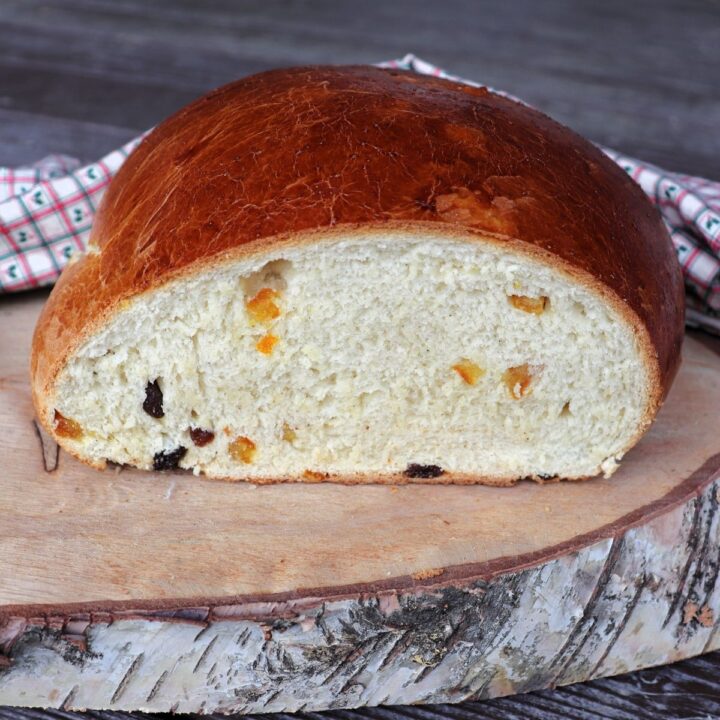 Slices of julekake bread sitting on a board next to the remaining loaf with bits of dried fruit exposed in the bread. A cloth sits behind it.