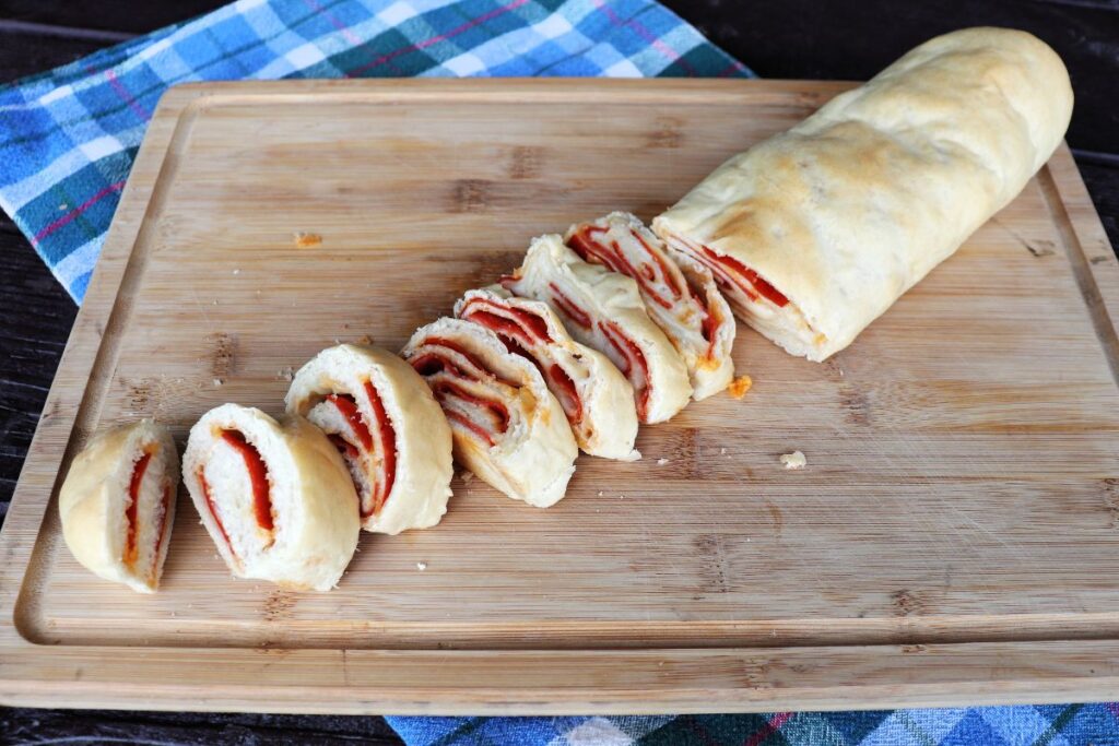 A loaf of pepperoni bread that has been partially sliced sitting on a cutting board that is sitting on blue checkered cloth.