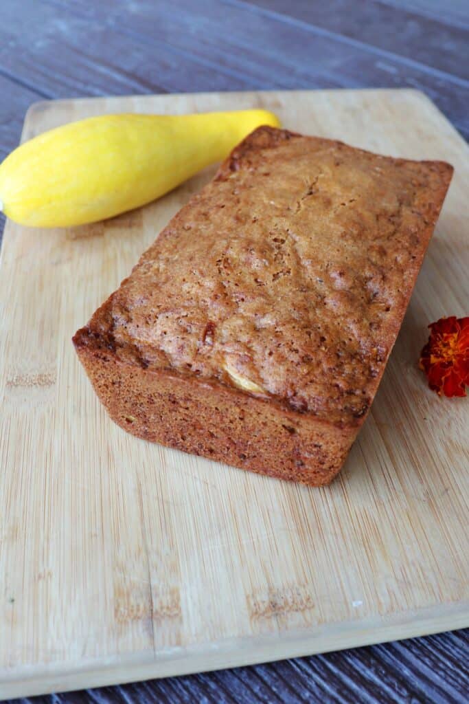 A loaf of squash bread sitting on a board with a yellow squash sitting behind it.