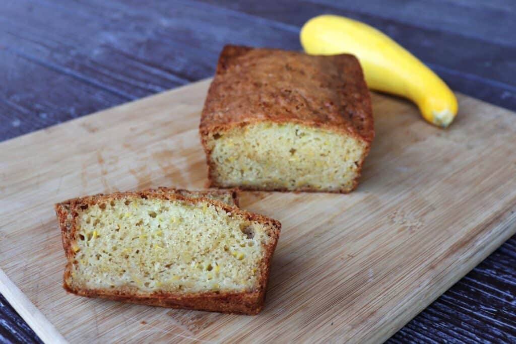 Slices of squash bread sitting on a board with remaining loaf behind it and a yellow summer squash.