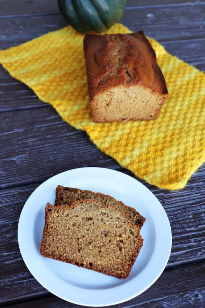 Slices of bread sit on a white plate. The remaining loaf sits on a yellow cloth behind it with a whole acorn squash.