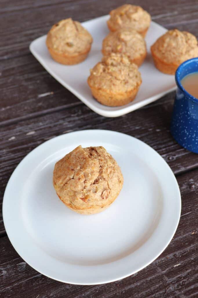 A single spelt muffin on a white plate as seen from above. A platter of more muffins sits behind it.