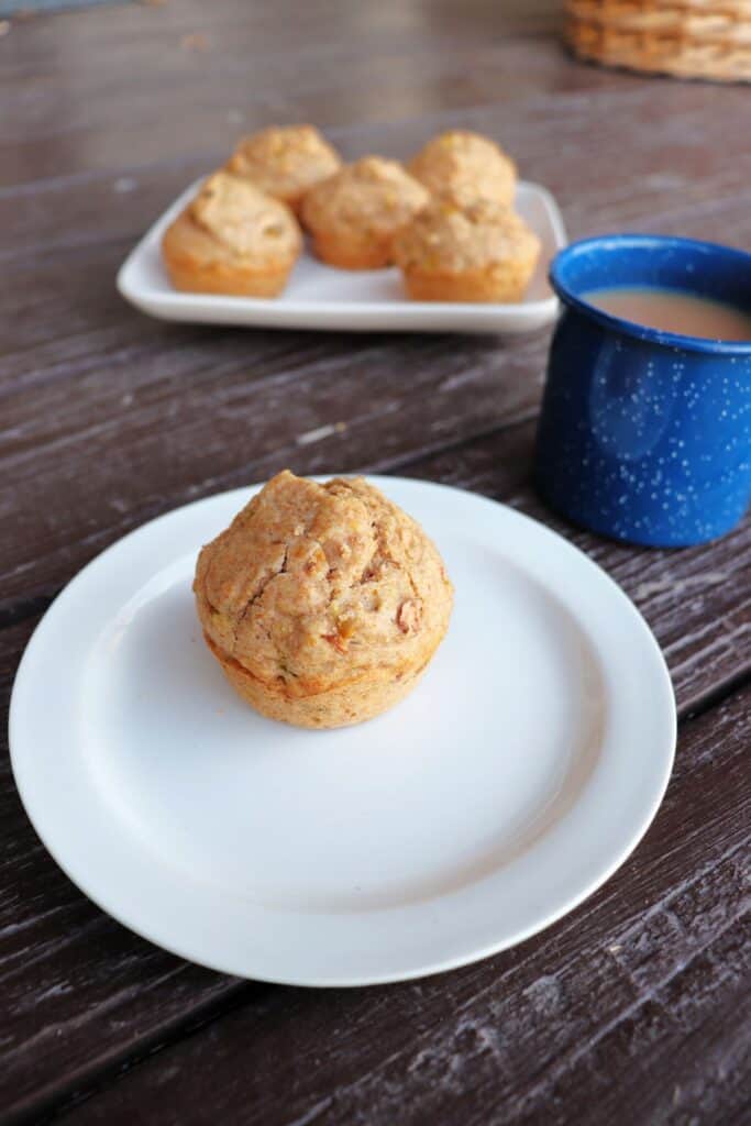 A single spelt muffin sits on a white plate. A blue tin cup with tea sits behind it. A platter of more muffins in the background.