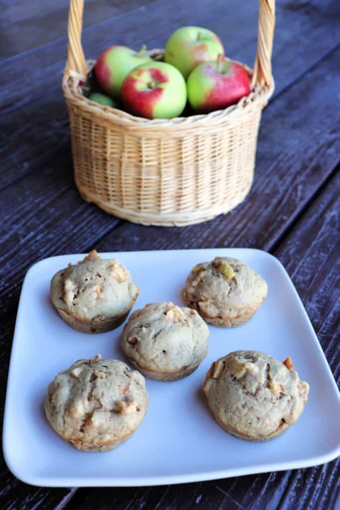 Apple spice muffins on a plate, a basket of fresh apples sits in the background.