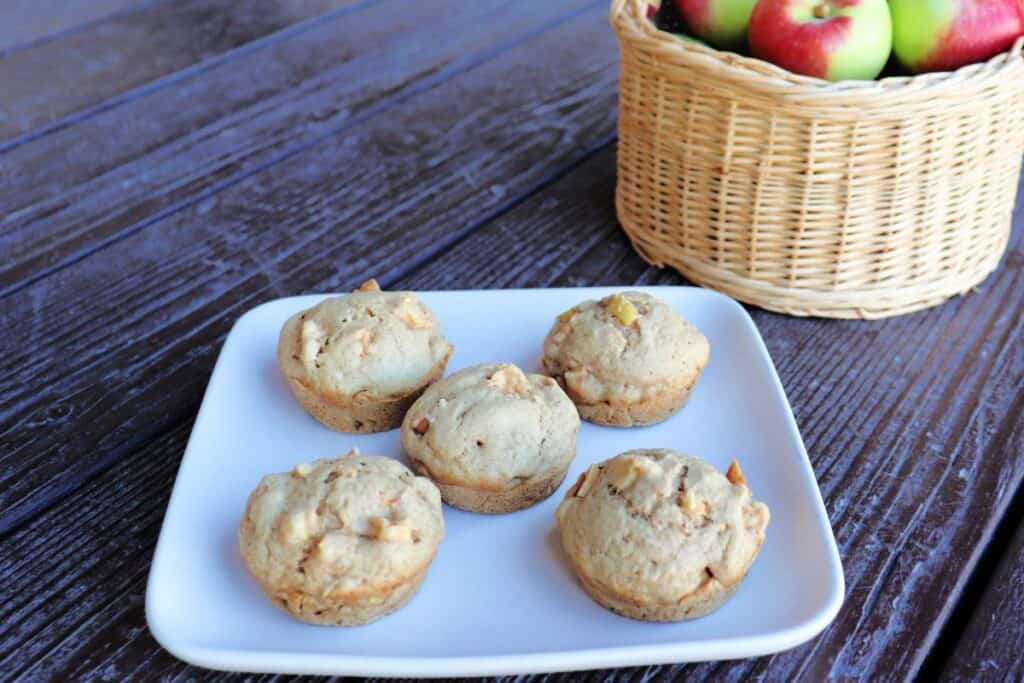 Apple spice muffins on a plate, a basket of fresh apples sits in the background.