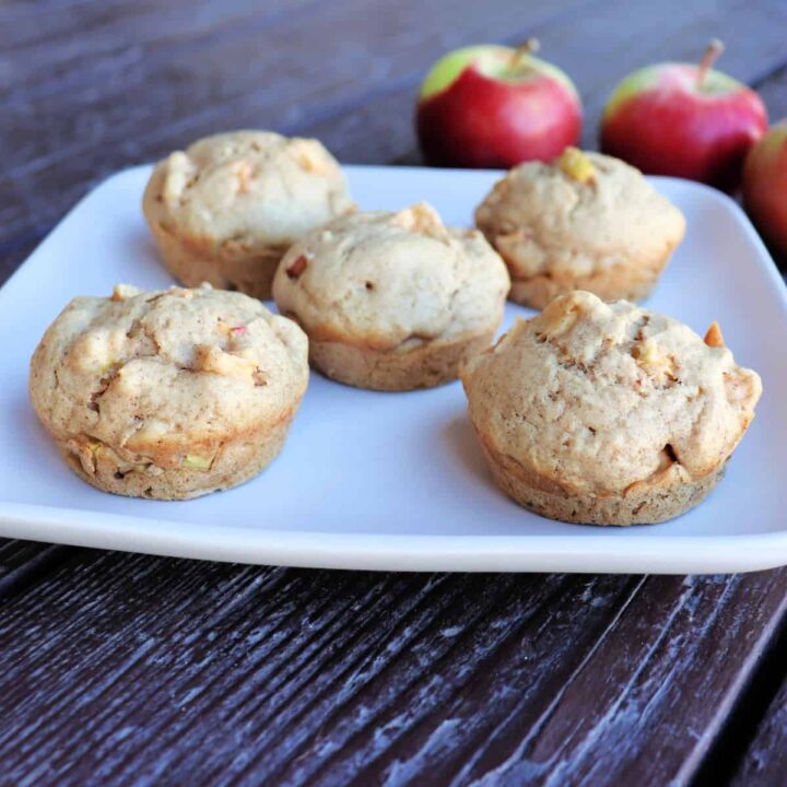 5 apple spice muffins sitting on a plate, fresh apples sit on the table behind it.