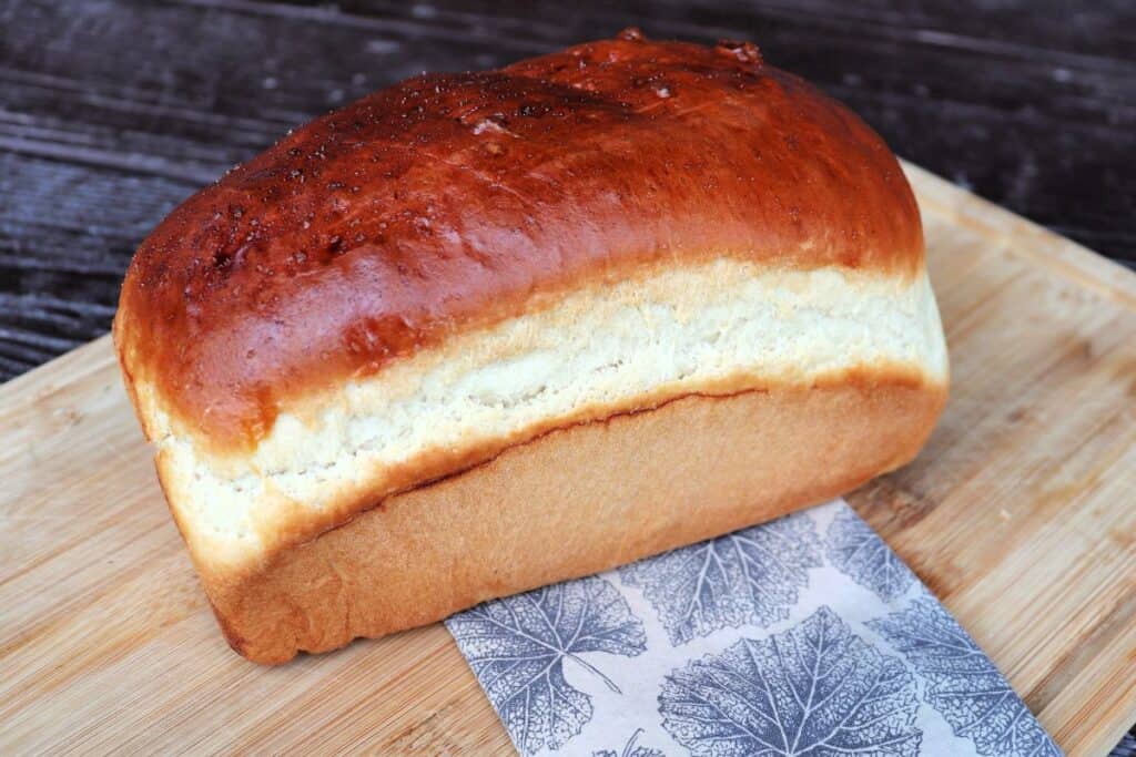 A golden loaf of white jamaican harddough bread sits on a cloth covered board.