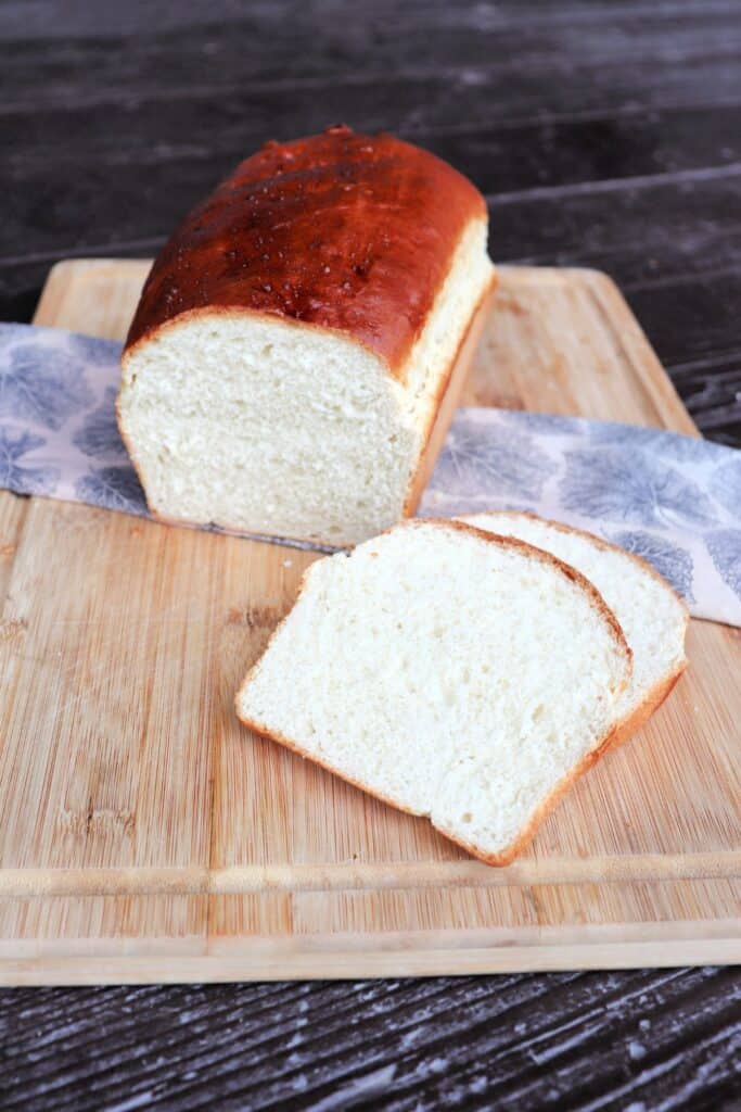 Slices of white bread sit on a board with remaining loaf siting on a cloth behind them. 