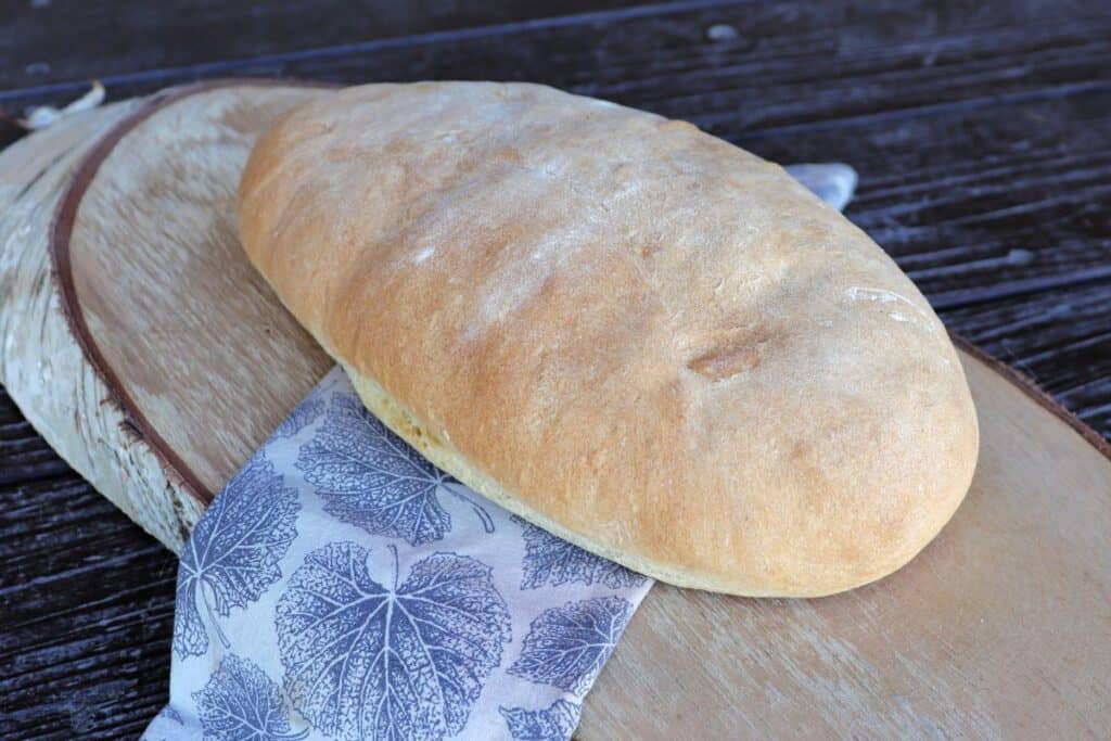 An oblong loaf of white hearth bread sits on a napkin that is draped over a wooden board as seen from the side.